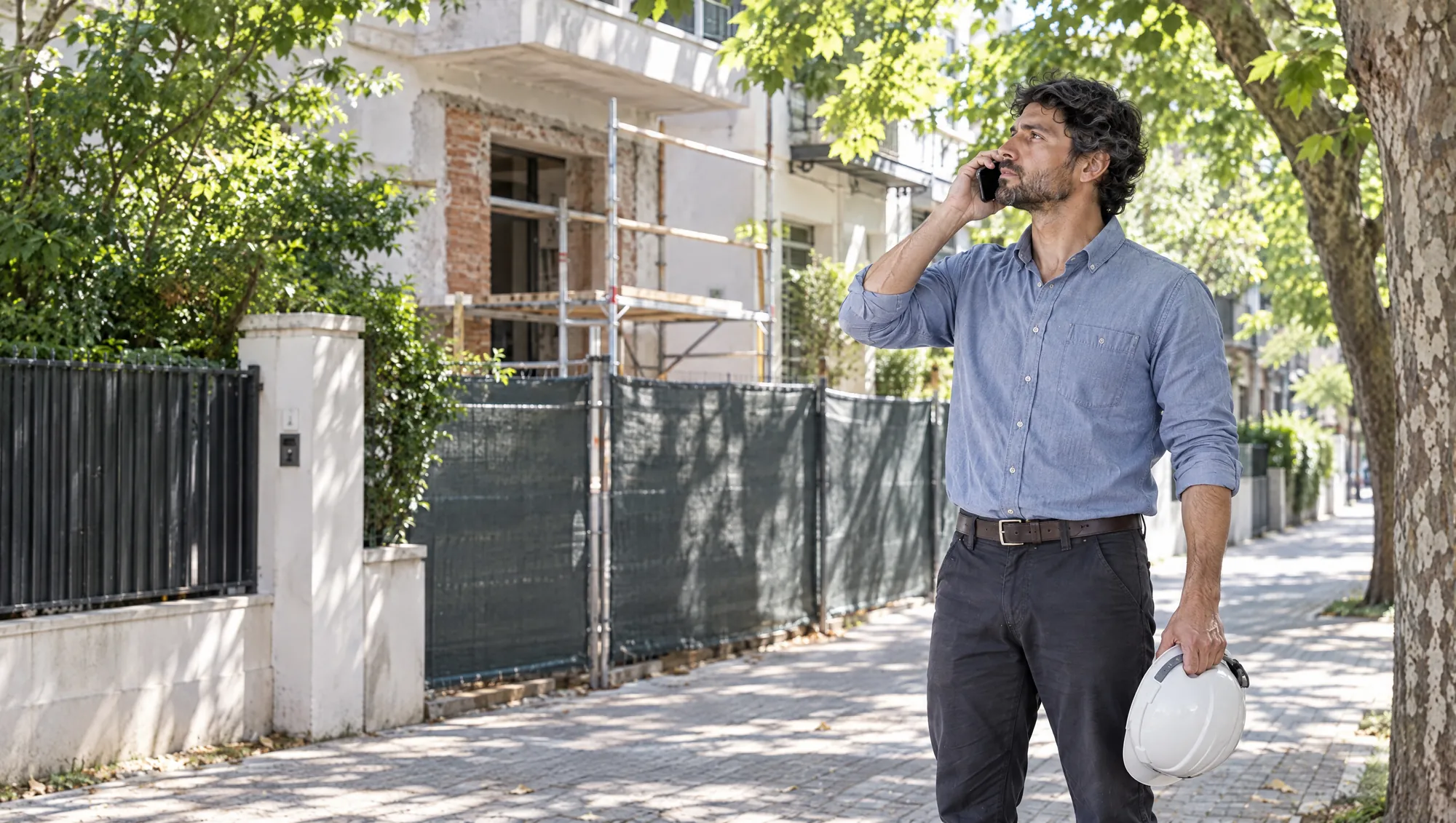 El Arquitecto de Estudio TREO atiende una consulta por celular en la vereda, frente a una casa en obra, con el casco en la mano
